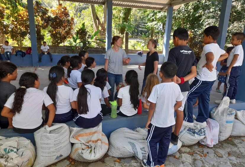 A volunteer talking with a group of schoolchildren in uniforms under an open-air shelter